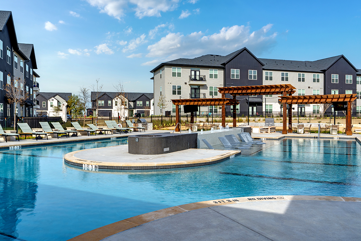 A modern apartment complex with a large outdoor swimming pool, sun loungers, wooden pergolas, and clear blue sky.
