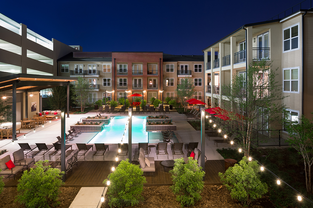 Modern apartment complex courtyard at night with a lit swimming pool, lounge chairs, red umbrellas, string lights, and landscaping, surrounded by multi-story buildings and a parking garage.