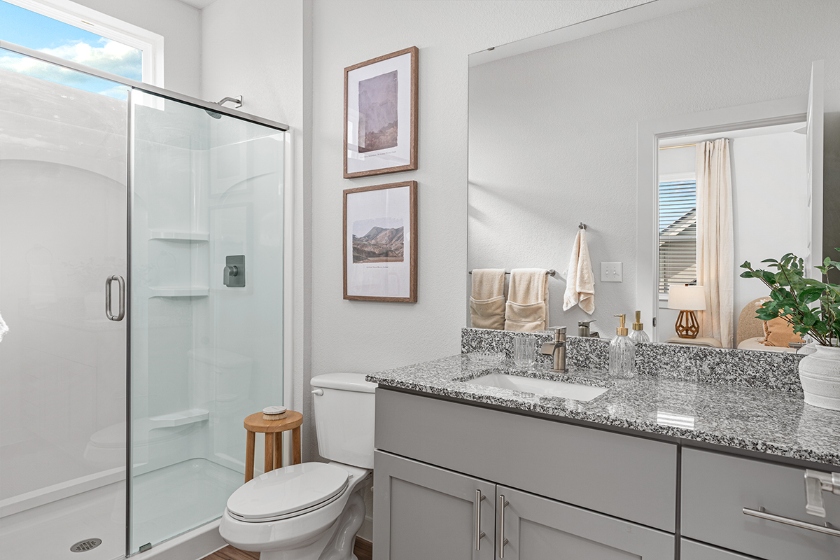 Modern bathroom with a glass shower, toilet, wooden stool, granite countertop, towels, potted plant, and framed art on white walls. A window lets in natural light.