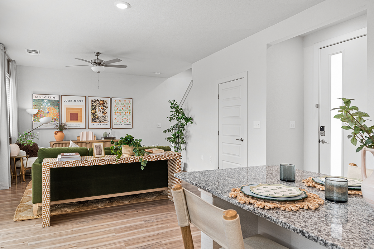 A bright, modern living room with a green sofa, wall art, indoor plants, wooden floor, and a kitchen island with placemats and glasses in the foreground. A door and white walls complete the space.