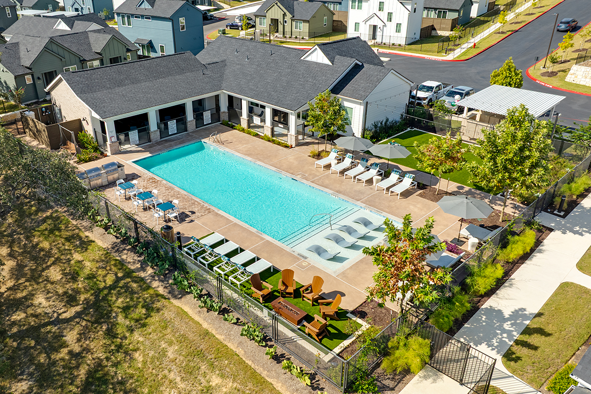 Aerial view of a modern apartment complex pool area with lounge chairs, umbrellas, shaded seating, a clubhouse, and landscaped surroundings on a sunny day.