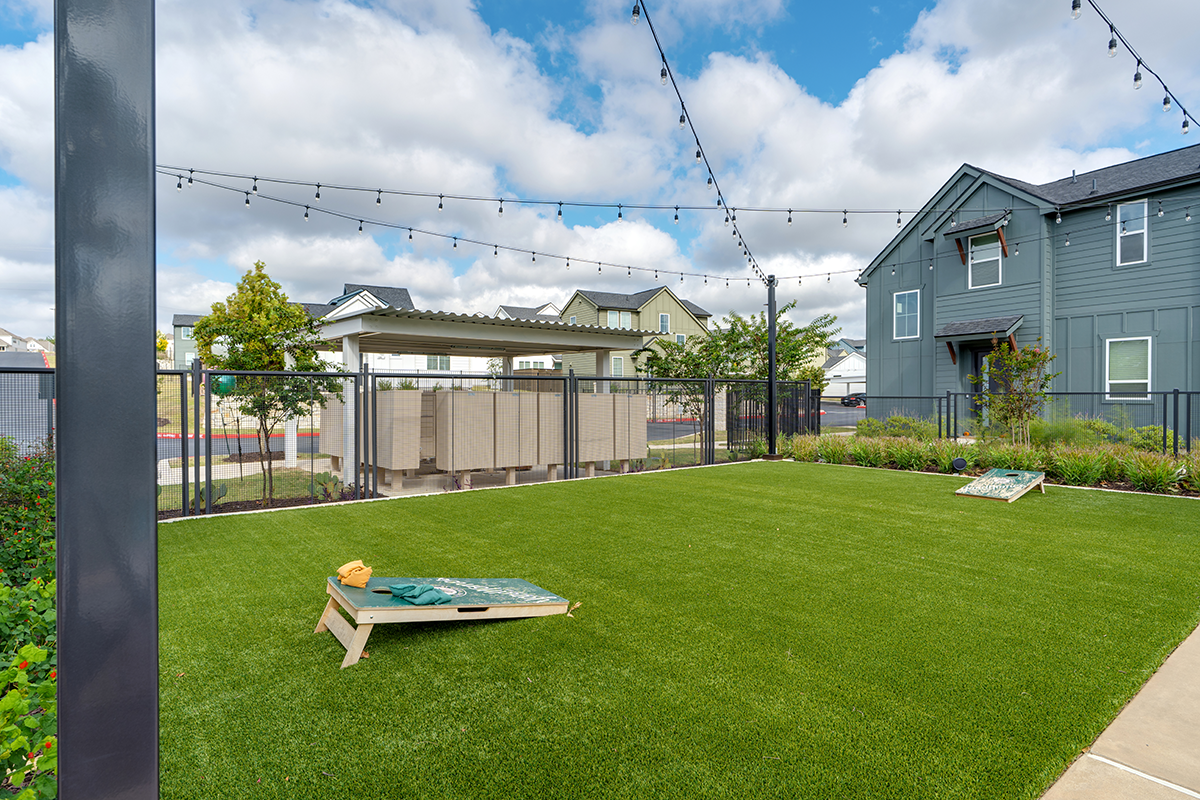 A backyard with artificial grass, string lights, two cornhole boards, and a covered seating area, surrounded by a black fence and houses in the background under a partly cloudy sky.