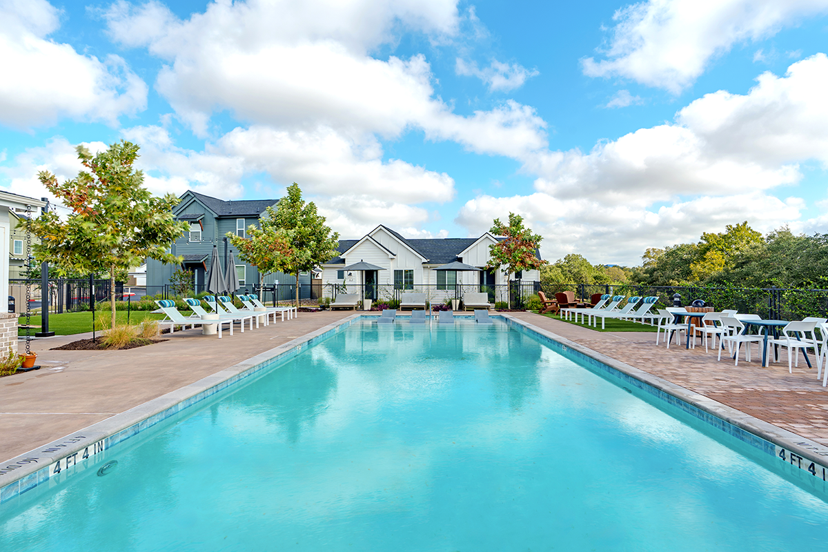 A clear outdoor swimming pool surrounded by lounge chairs, tables, and green trees, with residential buildings in the background under a partly cloudy blue sky.