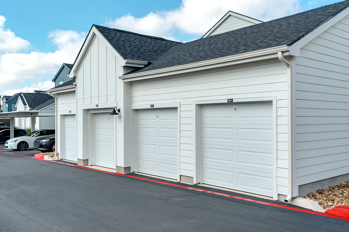 Row of three white, closed garage doors attached to a modern residential building with a dark roof, next to a paved driveway and parked cars on a sunny day.