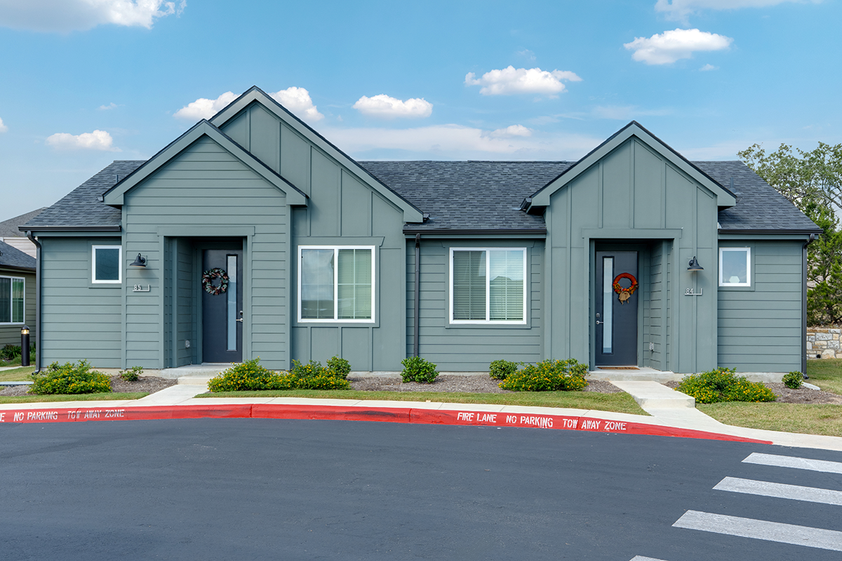 Two identical gray duplex homes with front doors and windows, small bushes along the walkway, and a red curb marked FIRE LANE NO PARKING TOW AWAY ZONE at the edge of a street under a partly cloudy sky.