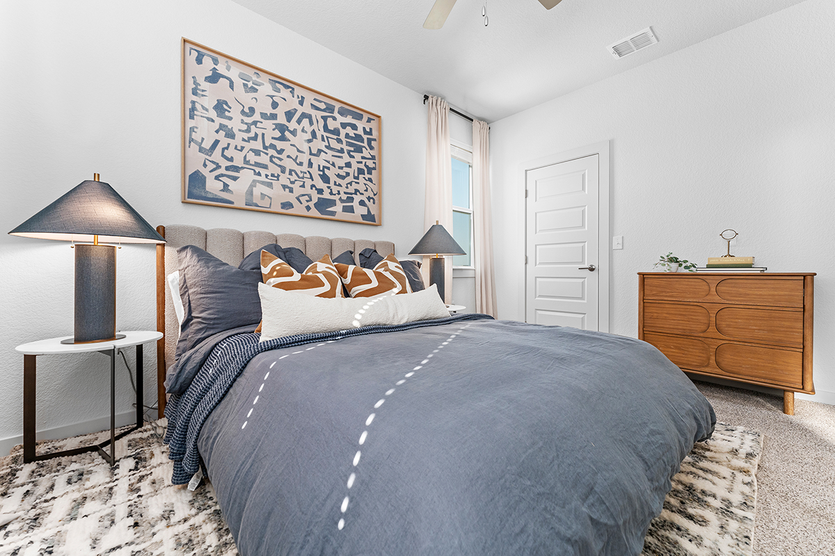 A modern bedroom with a gray bedspread, patterned pillows, two bedside lamps, abstract wall art, a wooden dresser, a window with light curtains, and a geometric rug.