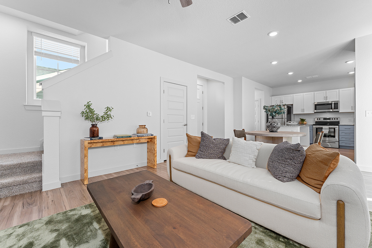 Modern living room with a white sofa, brown and gray cushions, wooden coffee table, and kitchen in the background. Stairs are to the left, and a console table with plants and decor sits against the wall.