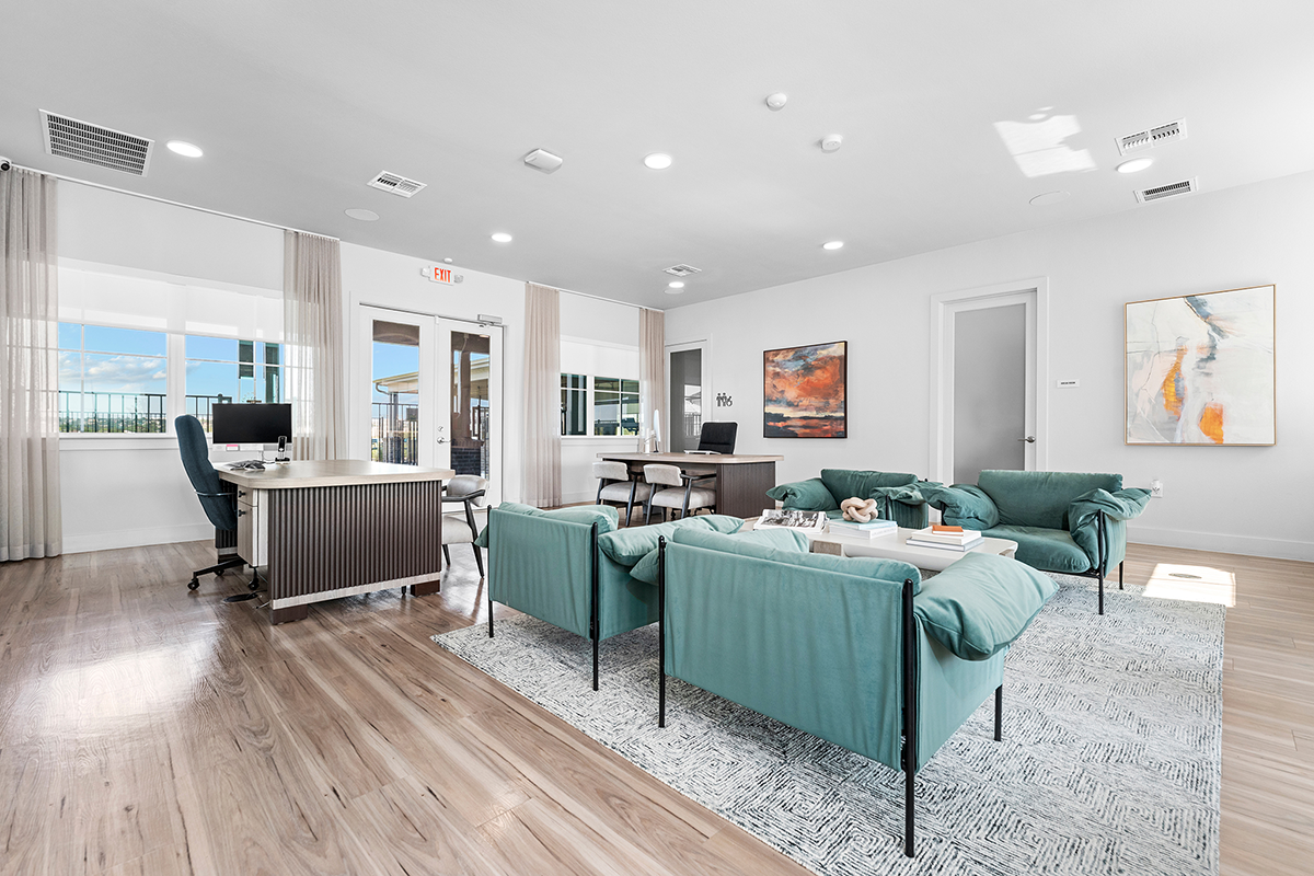 Modern office space with teal armchairs, a rug, coffee tables, a desk with a computer, large windows, white walls, wood flooring, and abstract artwork on the walls.