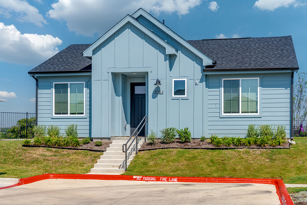 A small, light blue house with a dark roof, white trim, and front steps. There are two large windows and landscaping in front. A red-painted curb reads NO PARKING FIRE LANE. The sky is blue with scattered clouds.