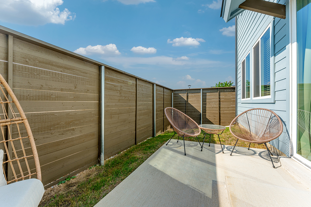 Small backyard patio with two modern wire chairs and a table on a concrete slab, enclosed by a tall wooden fence, next to a blue house on a sunny day with a few clouds.