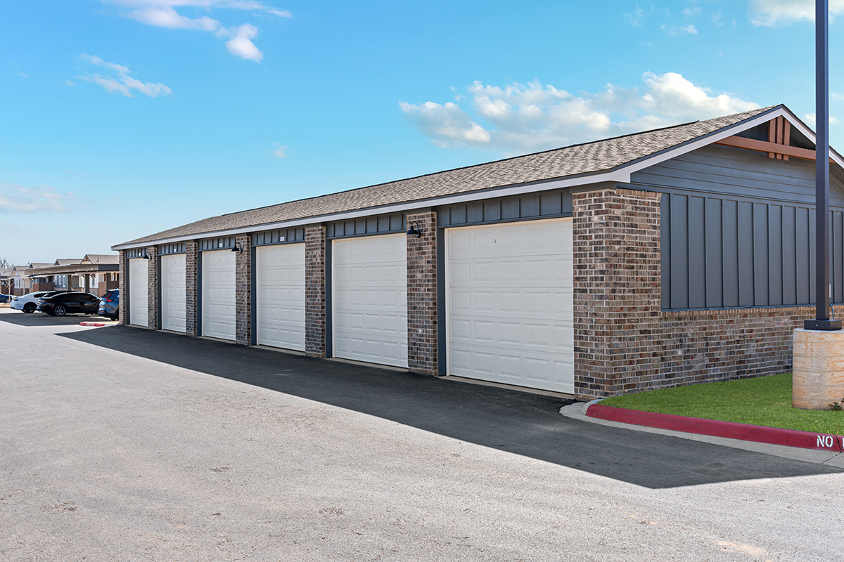 A row of six closed garage doors in a brick building next to a paved driveway, with blue sky and clouds overhead. Cars are parked in the background on the left side.
