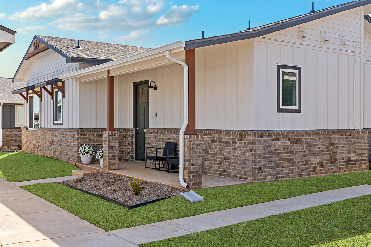 Modern single-story house with white siding and brick exterior, small covered front porch with two black chairs, potted flowers, and a neat lawn under a partly cloudy sky.