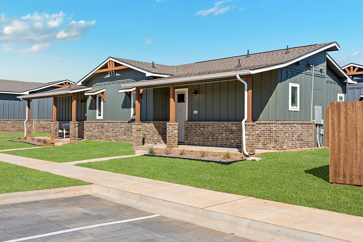A modern single-story house with gray siding, brick accents, and a neatly mowed lawn. The home has white trim, a small front porch, and is set near a concrete parking area under a clear blue sky.