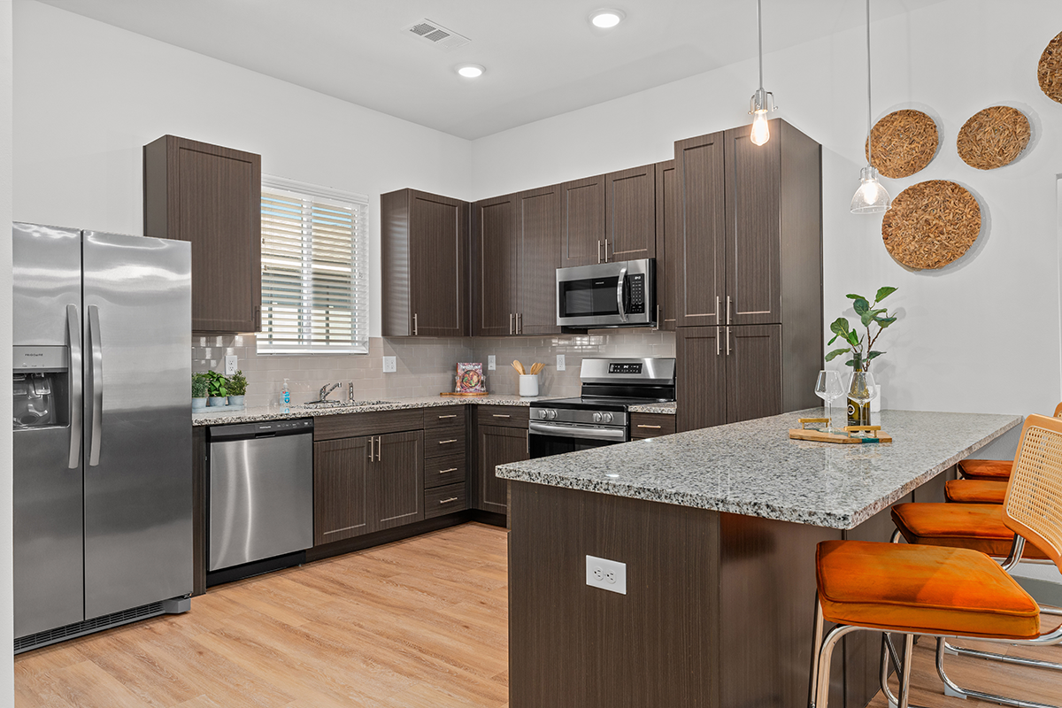 Modern kitchen with dark wood cabinets, stainless steel appliances, granite countertops, an island with orange barstools, light wood flooring, wall art, and a small plant on the counter.