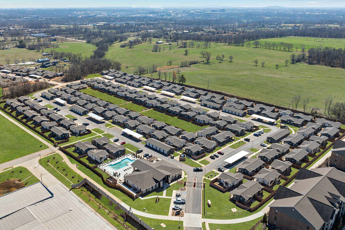 Aerial view of a suburban housing development with neatly arranged houses, streets, and a community pool, surrounded by open green fields and distant trees.