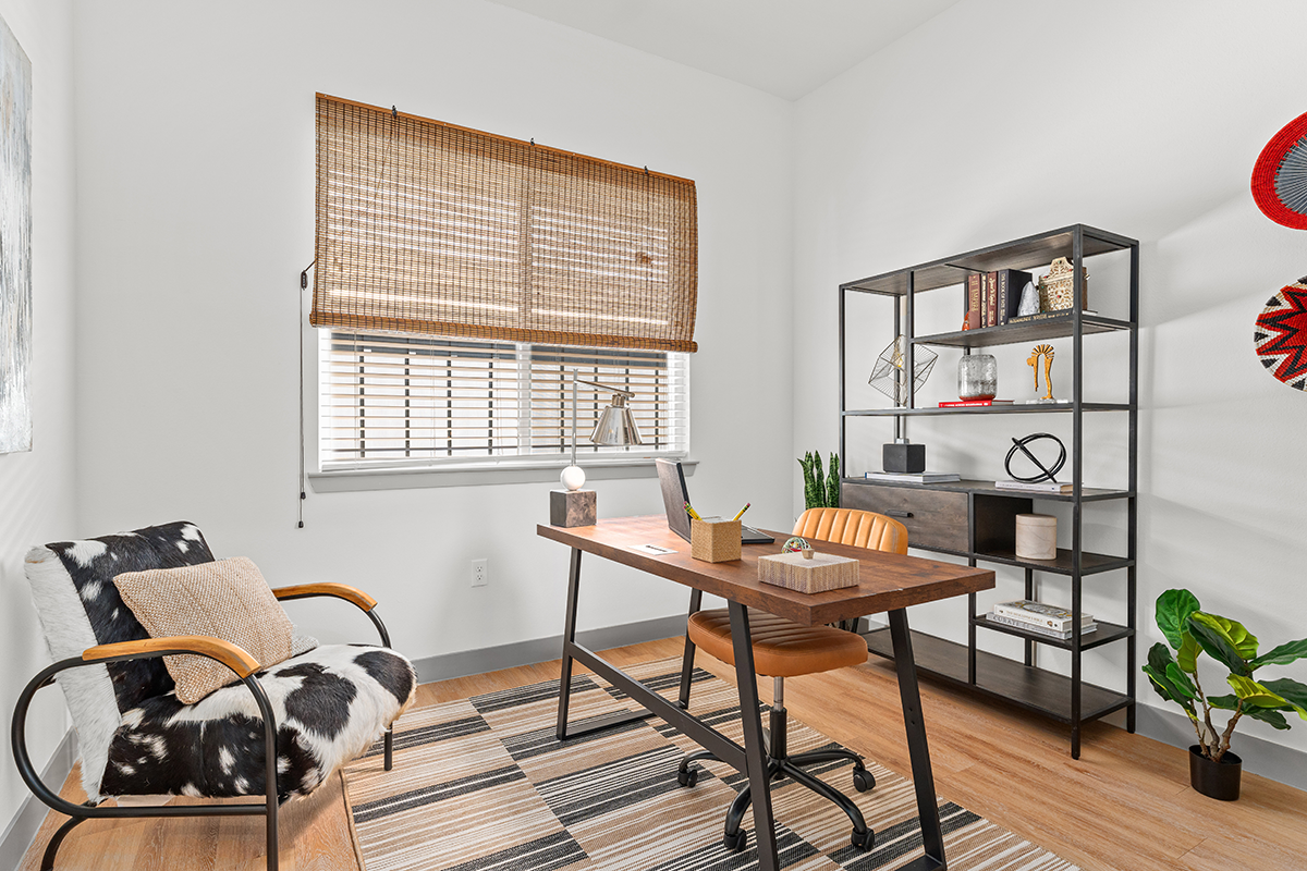 Modern home office with wooden desk, leather chair, cowhide accent chair, striped rug, open metal shelving, and a window with a bamboo shade. Decor includes plants, books, and geometric objects.