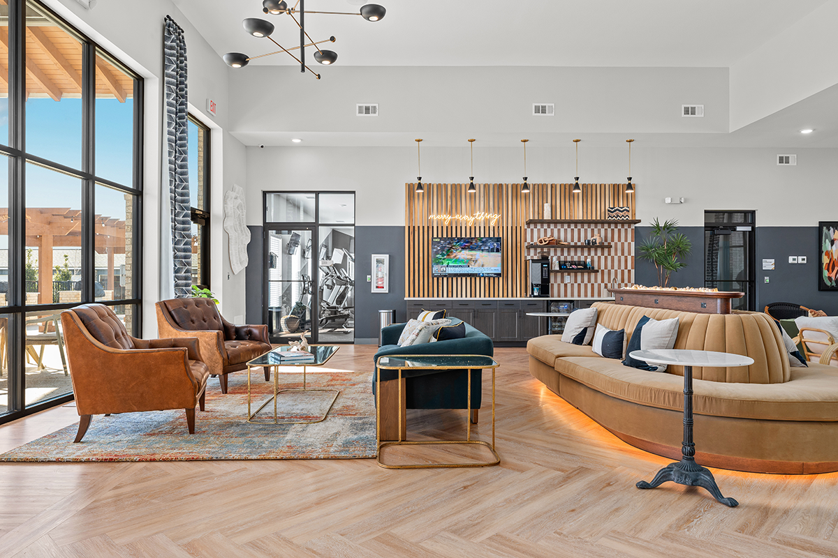 Modern lounge area with brown leather chairs, a curved beige sofa, coffee tables, wood flooring, large windows, indoor plants, and pendant lights. A kitchen or bar area with stools is visible in the background.