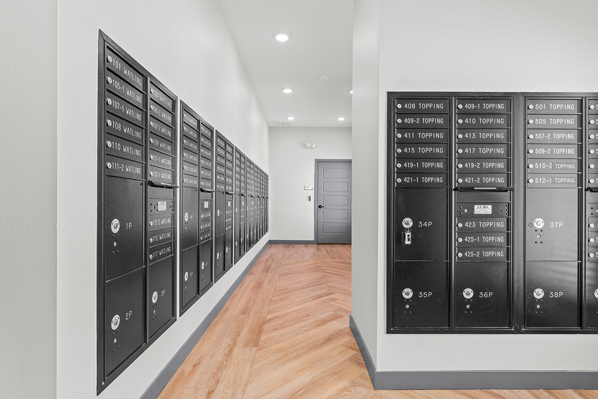 A hallway with black mailboxes lining both walls, featuring numbered boxes, bright lighting, and a wooden chevron-pattern floor leading to a gray door at the end.