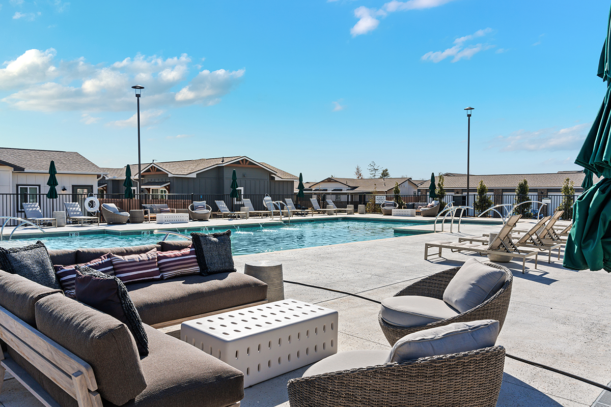 Outdoor pool area with lounge chairs, cushioned seating, umbrellas, and a clear blue sky above. Residential buildings are visible in the background.