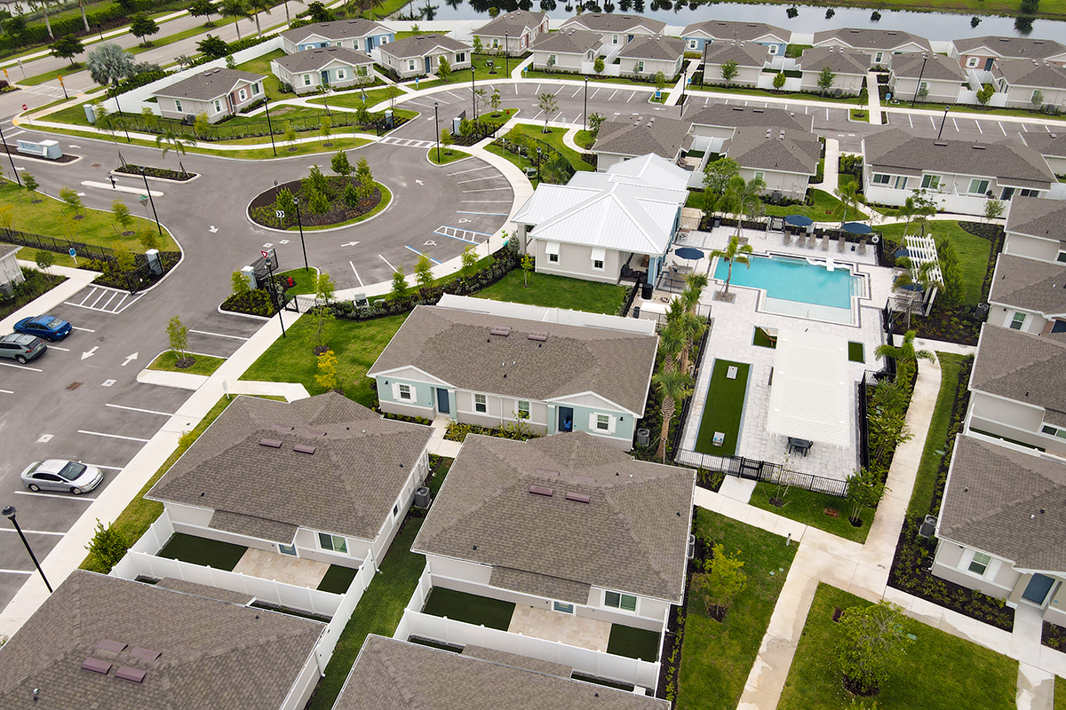 Aerial view of a suburban residential area with single-story houses, a swimming pool, parking lots, and landscaped green spaces. Pathways connect homes, and a pond is visible in the background.