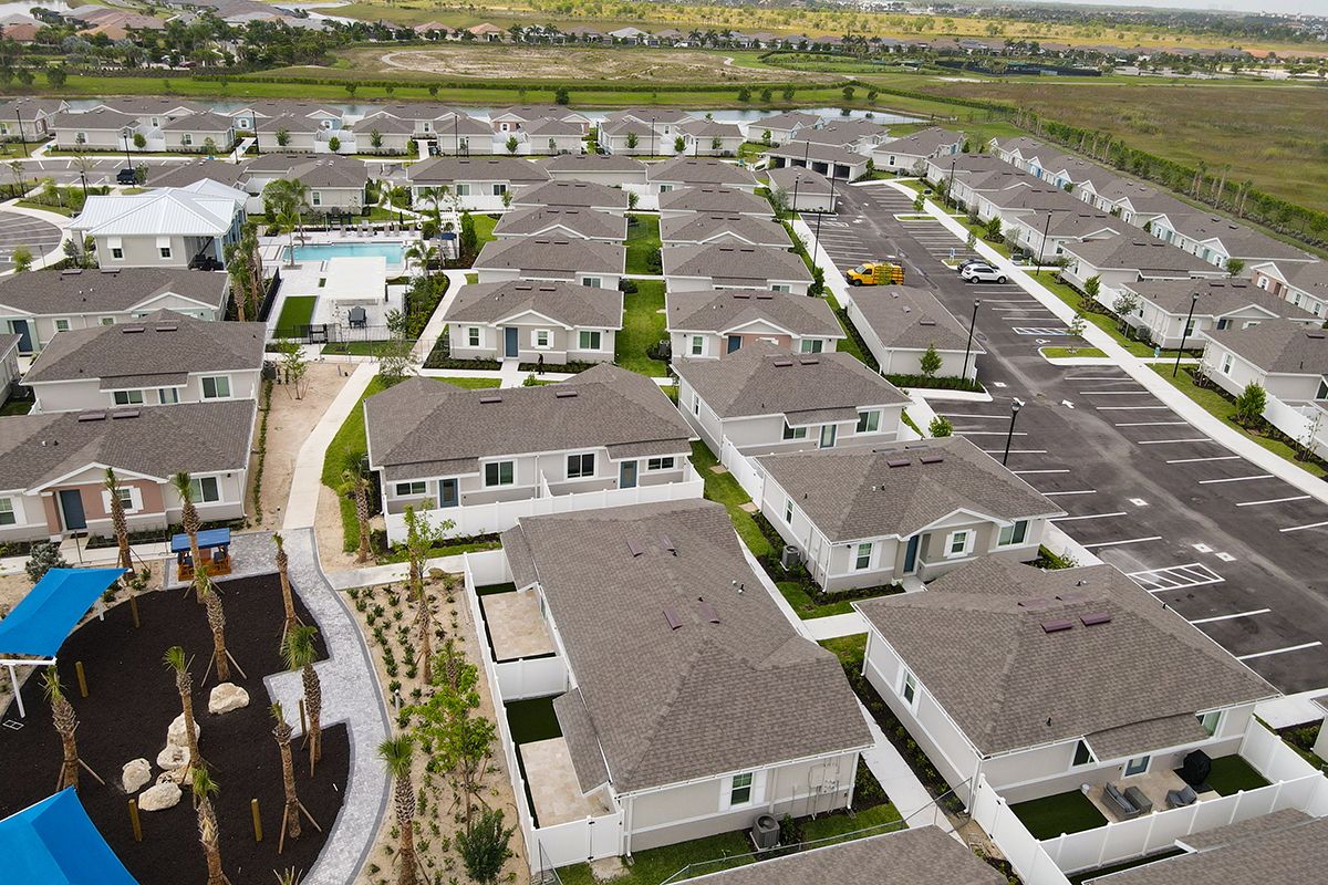 Aerial view of a suburban neighborhood with rows of single-story houses, a swimming pool, parking lots, a playground, landscaped areas, and open fields in the background.