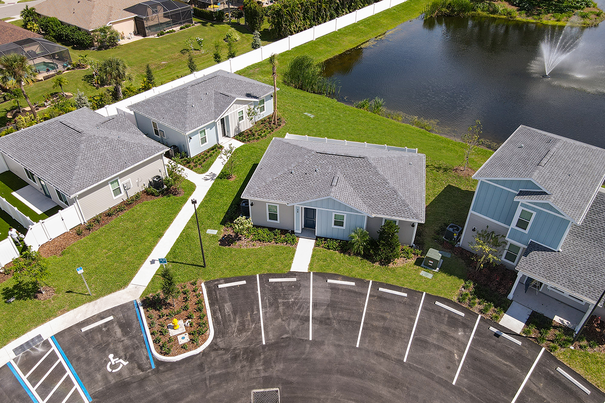 Aerial view of several small houses with gray roofs near a pond with a fountain, surrounded by green grass. Empty parking spaces, including a marked accessible spot, are visible in the foreground.