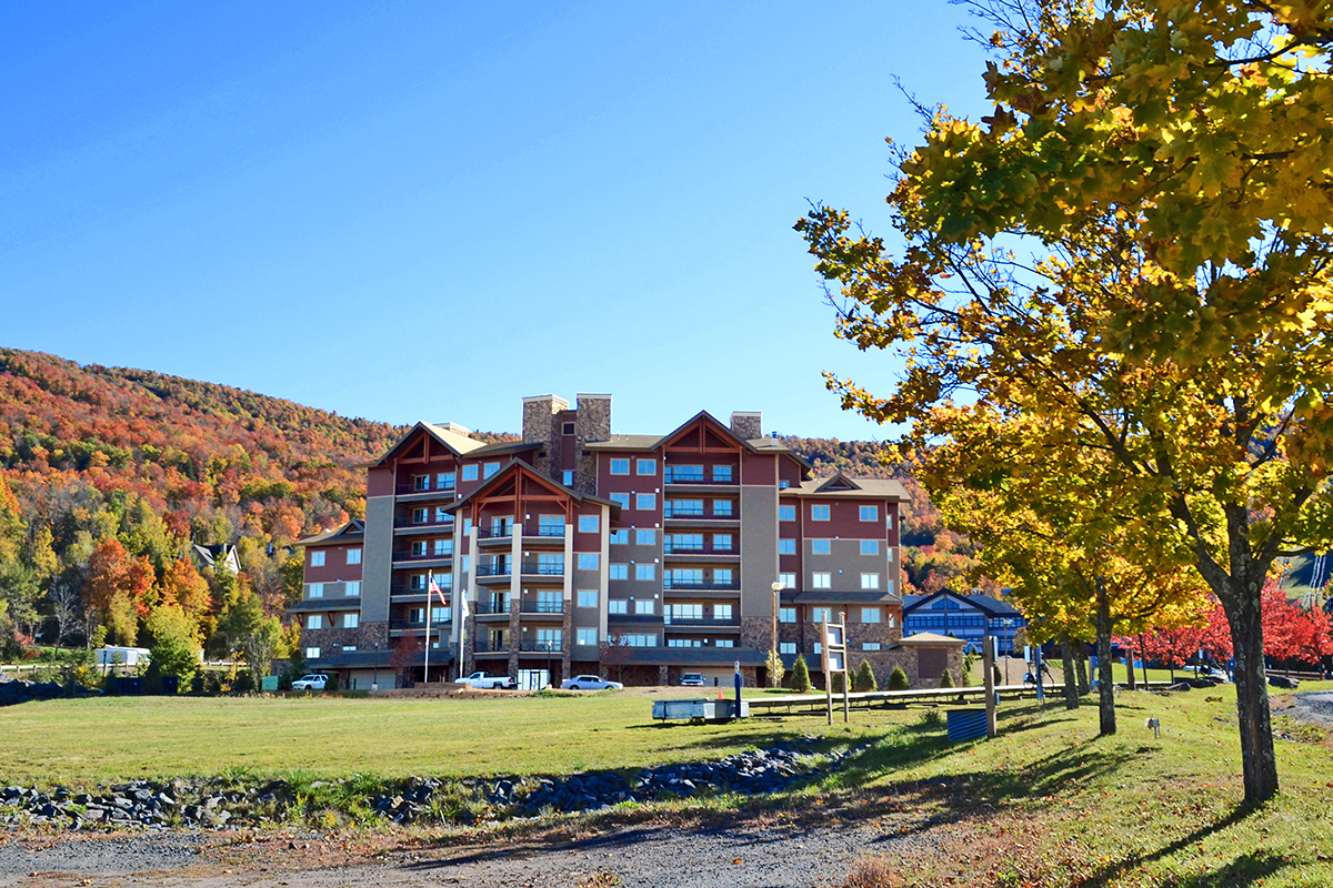 A lodge-style hotel with brown accents sits at the edge of a grassy field, surrounded by autumn trees and colorful hills under a clear blue sky.