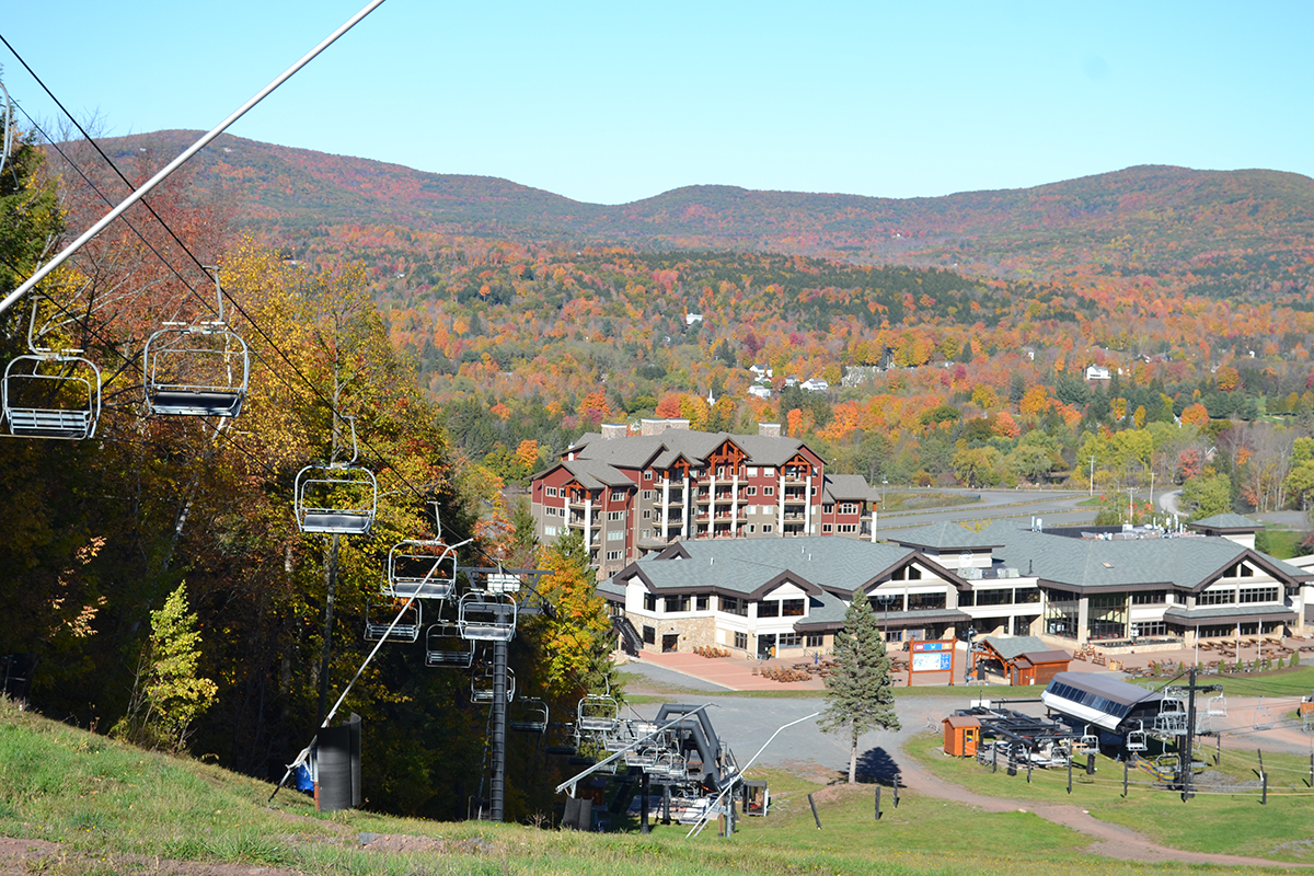Ski lifts and buildings at the base of a mountain, surrounded by colorful autumn trees and distant hills under a clear blue sky.