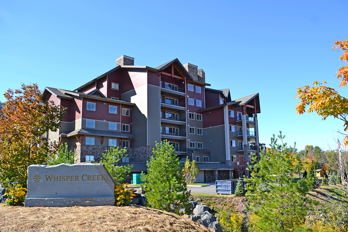 A multi-story lodge-style building with balconies, surrounded by trees and a stone sign in front that reads Whisper Creek under a clear blue sky.