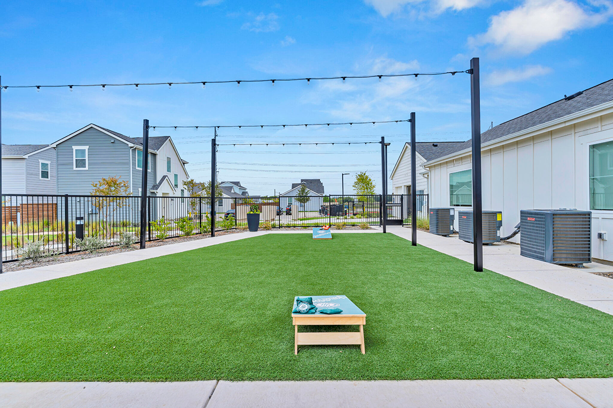Outdoor artificial turf area with cornhole boards, string lights overhead, modern houses, and air conditioning units along the right. The sky is blue with a few clouds.