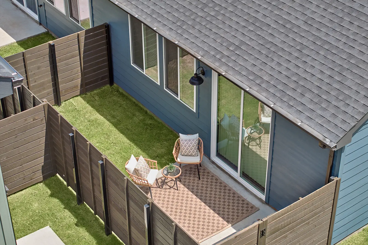 A small fenced backyard with green grass, a patterned rug, two chairs with pillows, and a small round table beside a blue house with large windows and a sliding glass door.