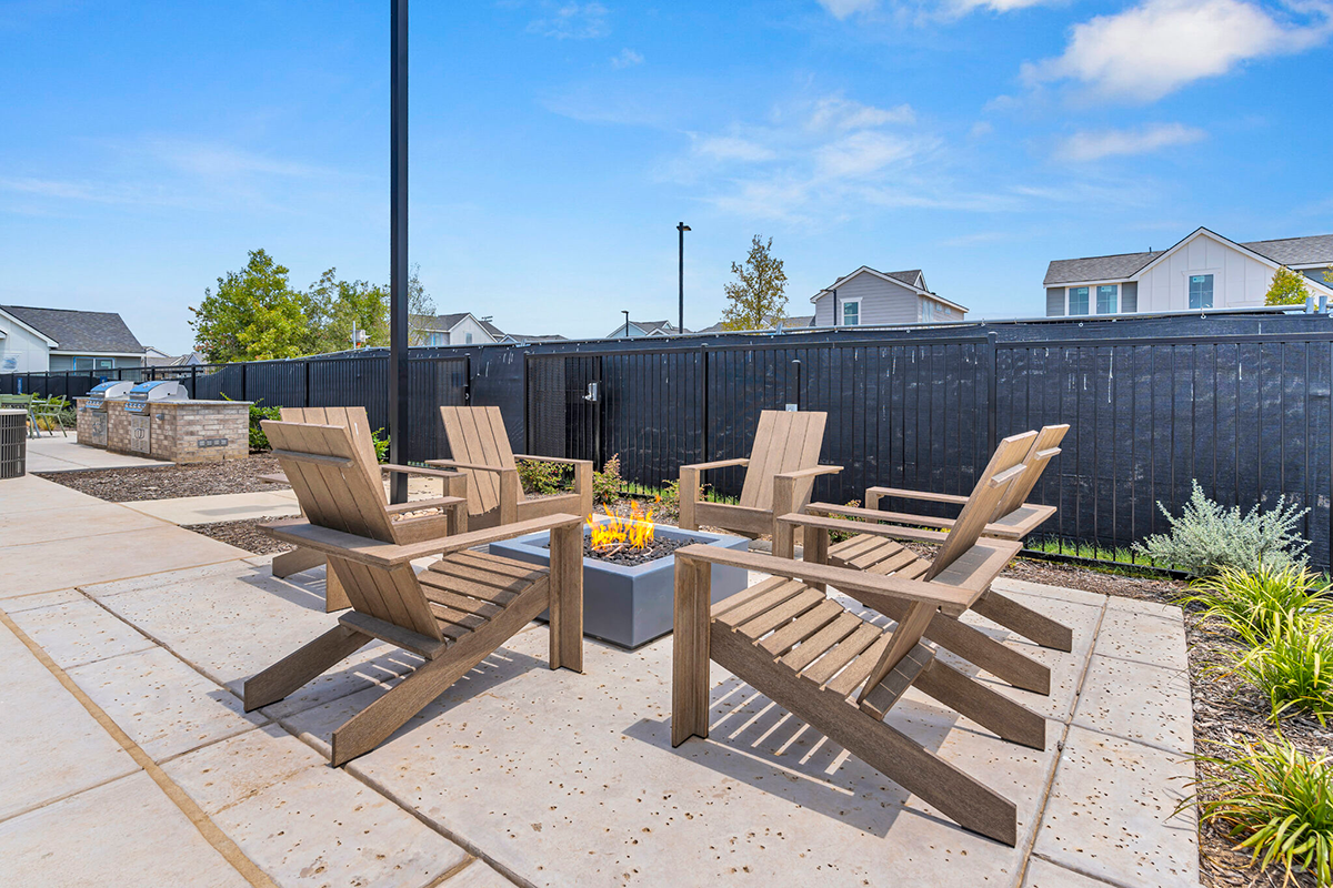 Four wooden Adirondack chairs surround a square fire pit on a patio, with a black fence and houses in the background under a blue sky.