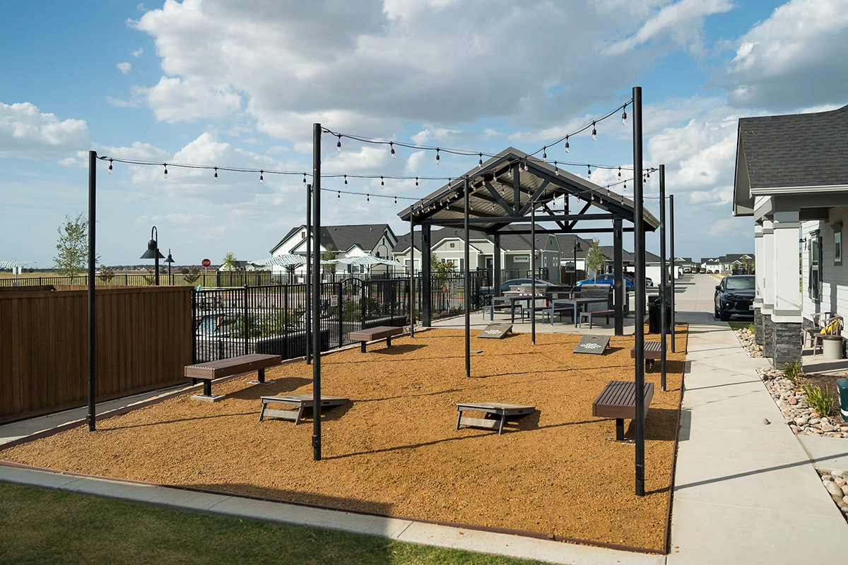 A covered outdoor seating area with benches and swings under string lights, surrounded by a gravel surface, next to a fenced yard and modern houses under a partly cloudy sky.