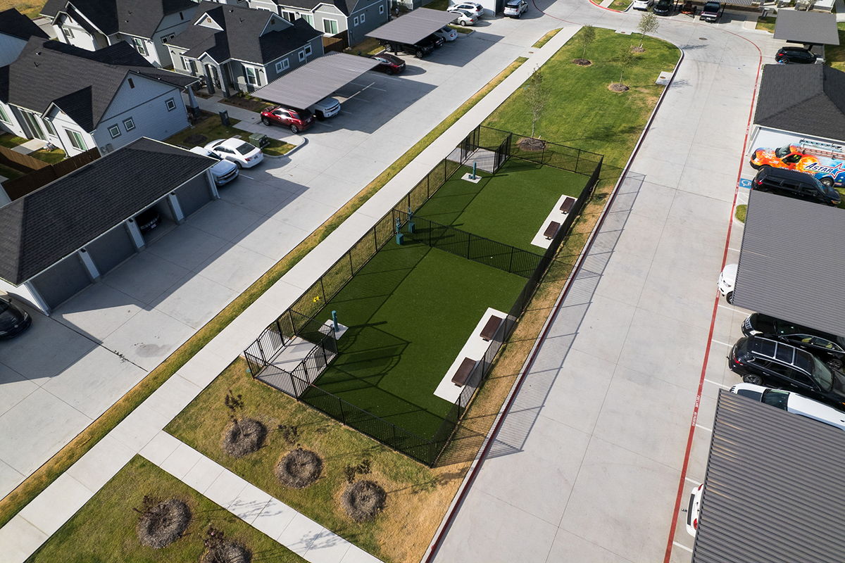 A fenced dog park with artificial grass, benches, and shaded areas, located between residential buildings and parking spaces in a suburban neighborhood.
