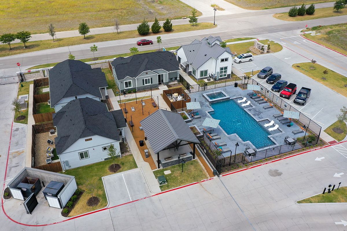Aerial view of a residential community with a central pool, lounge chairs, small houses, cars parked nearby, and fenced outdoor areas on a quiet street.