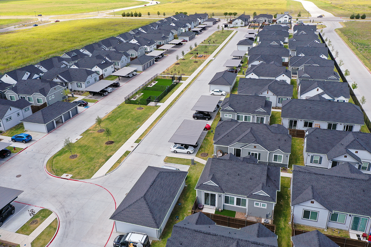 Aerial view of a suburban neighborhood with rows of single-story houses, driveways, parked cars, manicured lawns, and surrounding green fields.