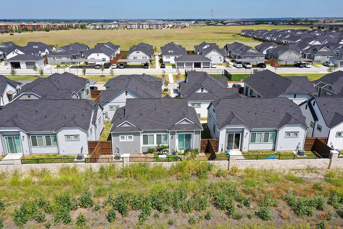 Aerial view of modern suburban houses with gray roofs, backyards, and green lawns, bordered by an open grassy area and a row of shrubs in the foreground.
