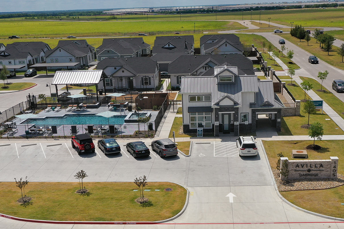 Aerial view of a residential complex with a swimming pool, parking lot with cars, lawn, and clubhouse. The sign reads AVILLA CAMBRIDGE. Fields and a road are visible in the background under a partly cloudy sky.