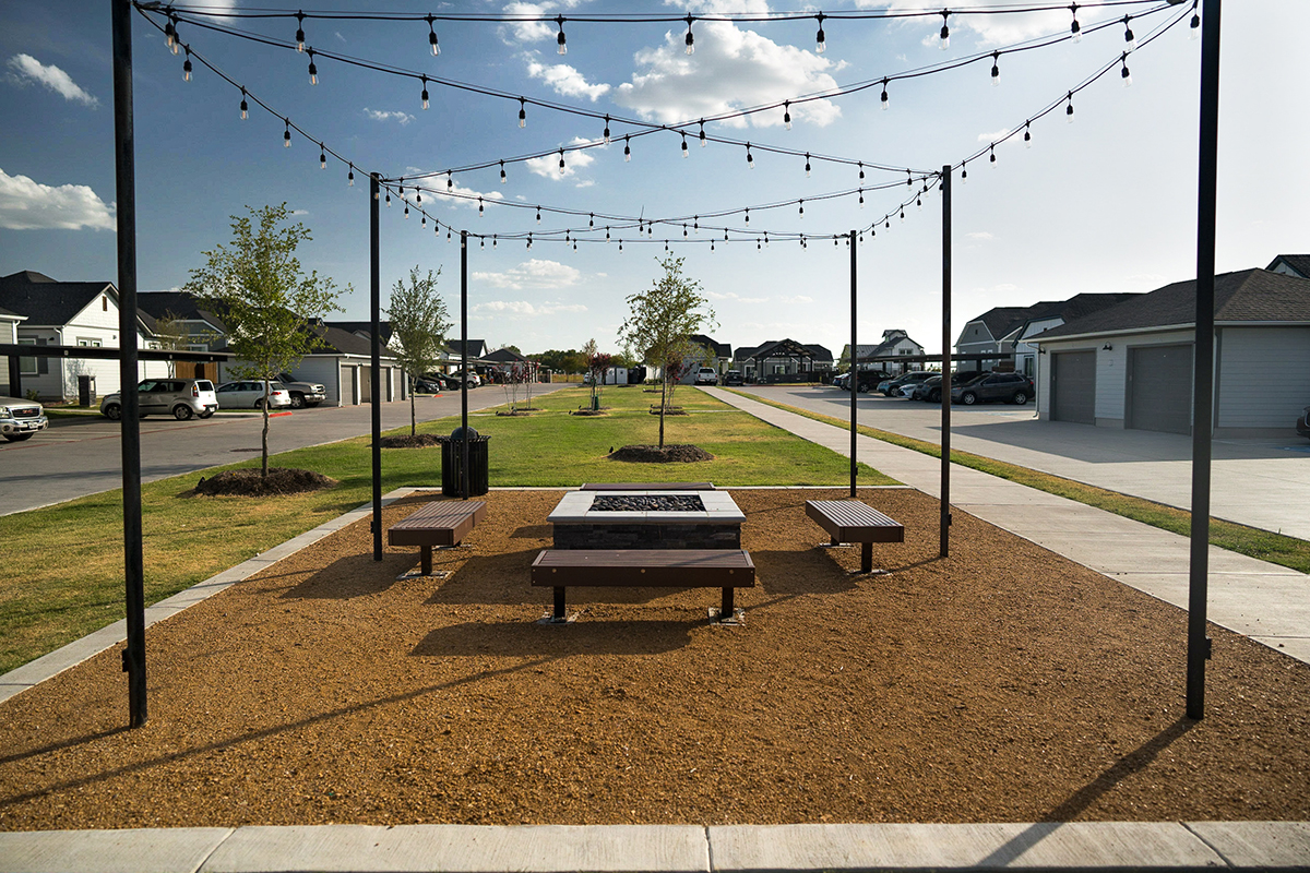 A small outdoor fire pit area with benches, string lights, and young trees in a residential neighborhood under a partly cloudy sky.