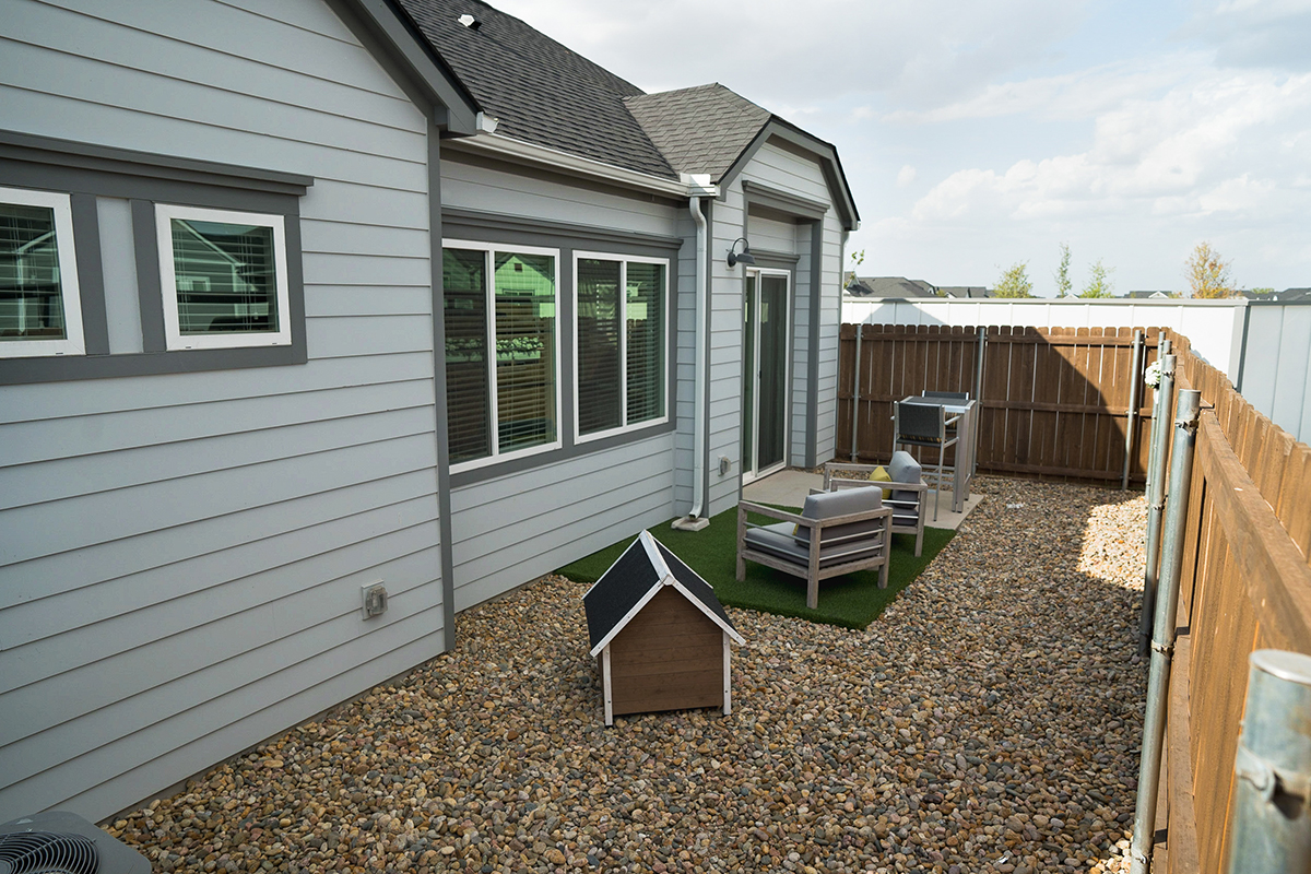 A small backyard with a gravel ground, two chairs and a table on artificial grass, a doghouse, and a wooden fence surrounding a light gray house with several windows.