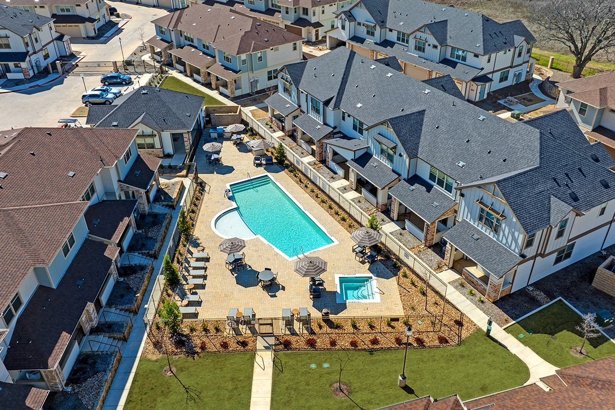 Aerial view of a modern apartment complex featuring a central outdoor swimming pool, lounge chairs, umbrellas, landscaped paths, and surrounding townhomes.