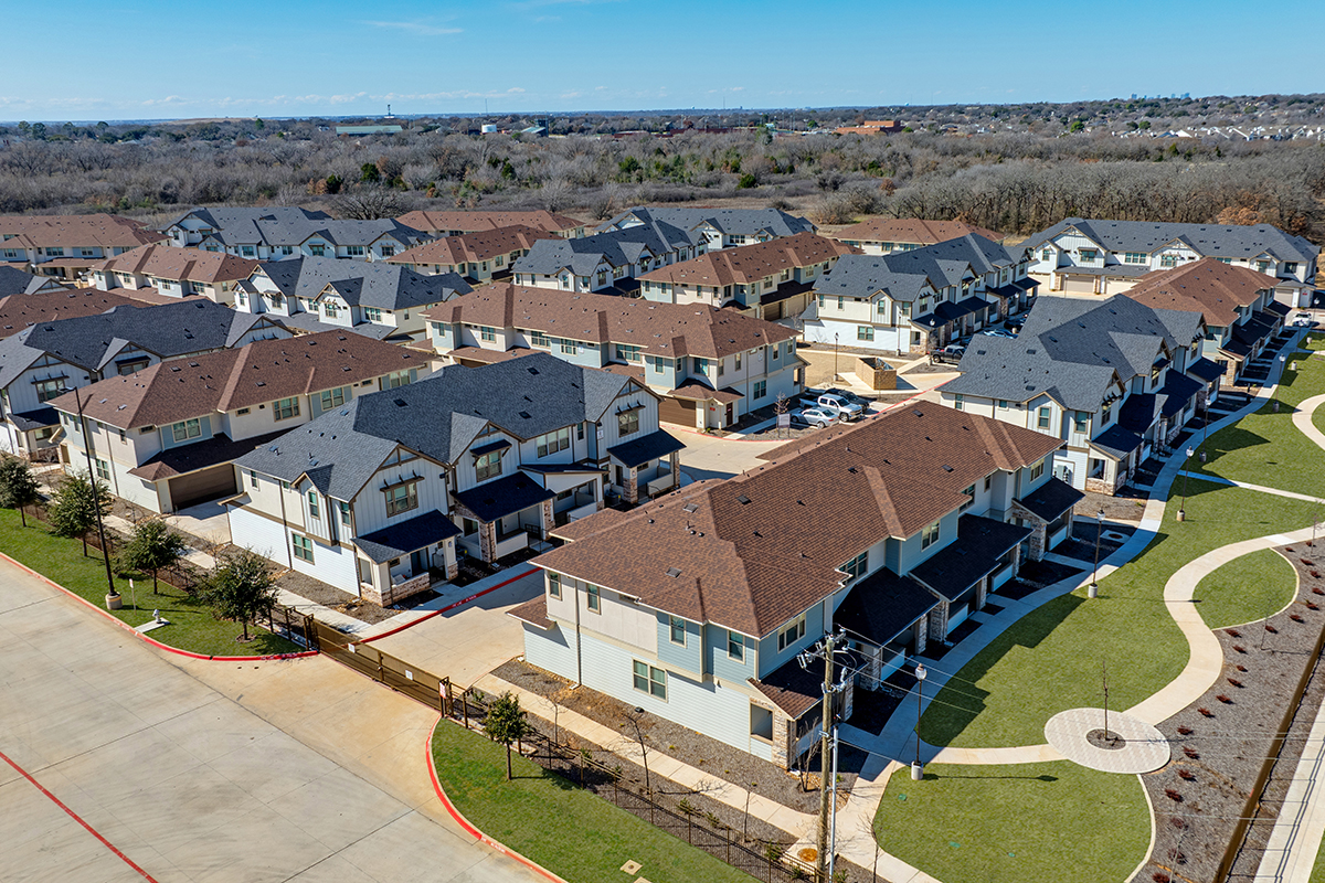 Aerial view of a suburban neighborhood showing rows of modern townhouses with gray and brown roofs, green lawns, and a curving sidewalk under a clear blue sky.