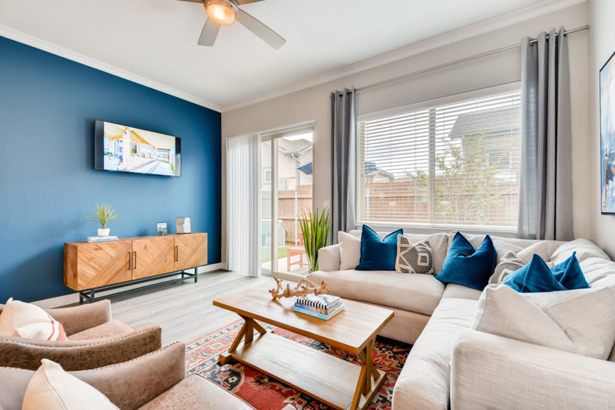 Bright living room with a sectional sofa, blue accent wall, TV, wooden coffee table, two armchairs, large window with blinds, and decorative pillows. Modern, cozy, and filled with natural light.