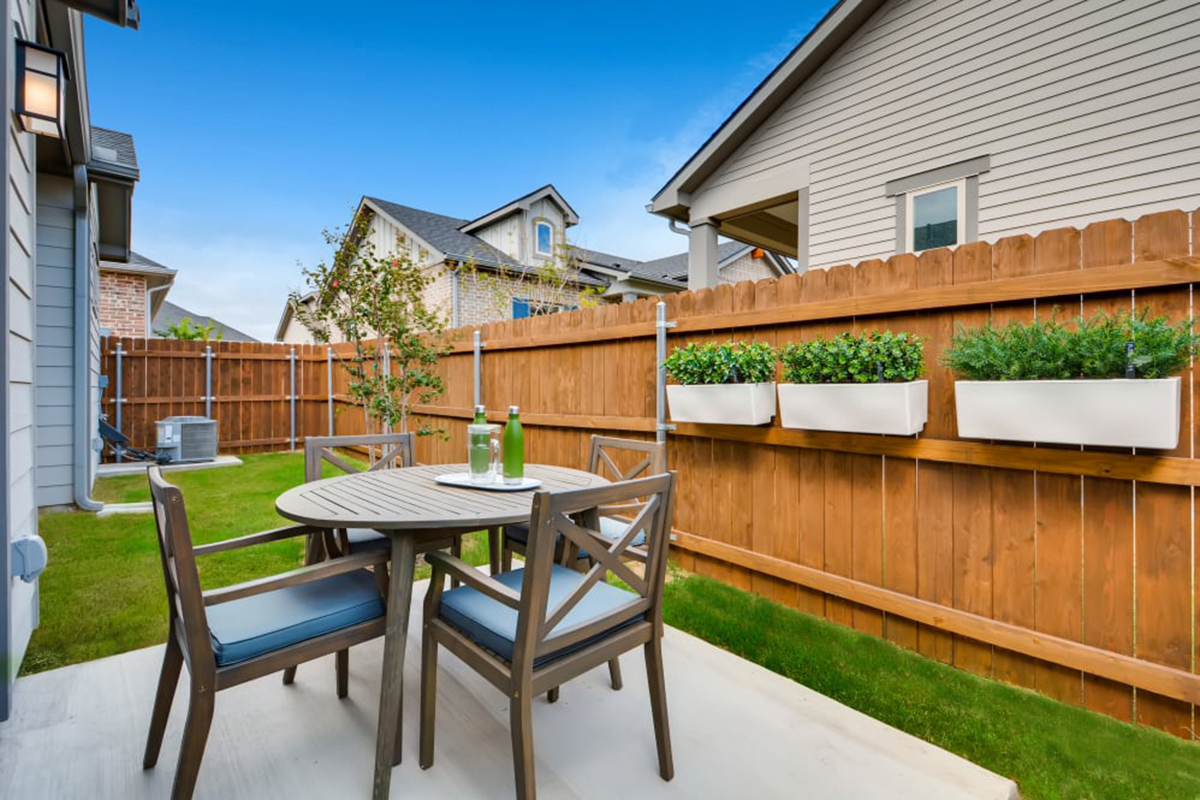 A small patio with a round table, four chairs, two bottles, and glasses. The yard has green grass, a wooden fence with planter boxes, and nearby houses under a blue sky.