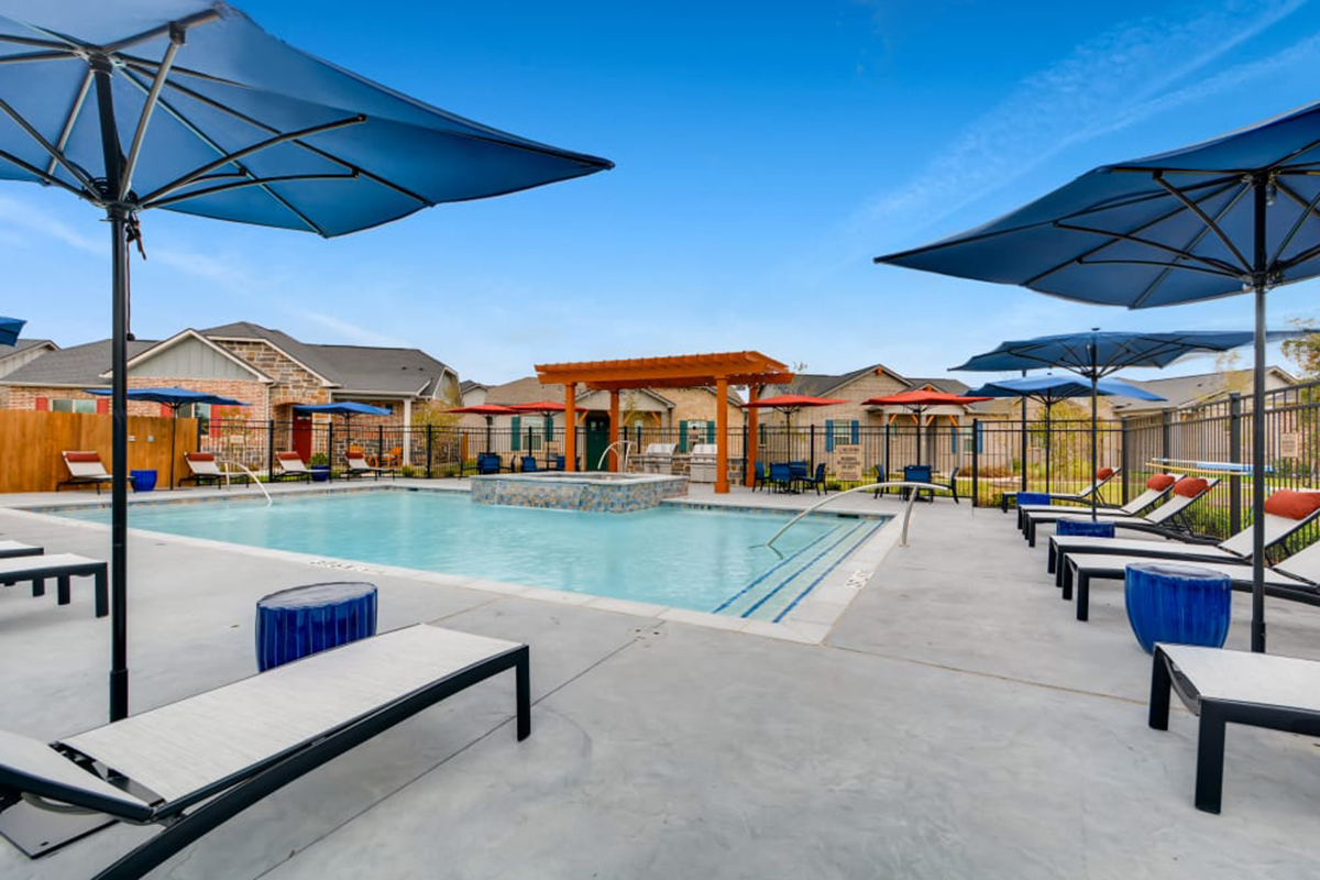 Outdoor swimming pool with lounge chairs, blue umbrellas, and a wooden pergola in the background. The sky is clear and blue, and houses are visible behind the pool area.