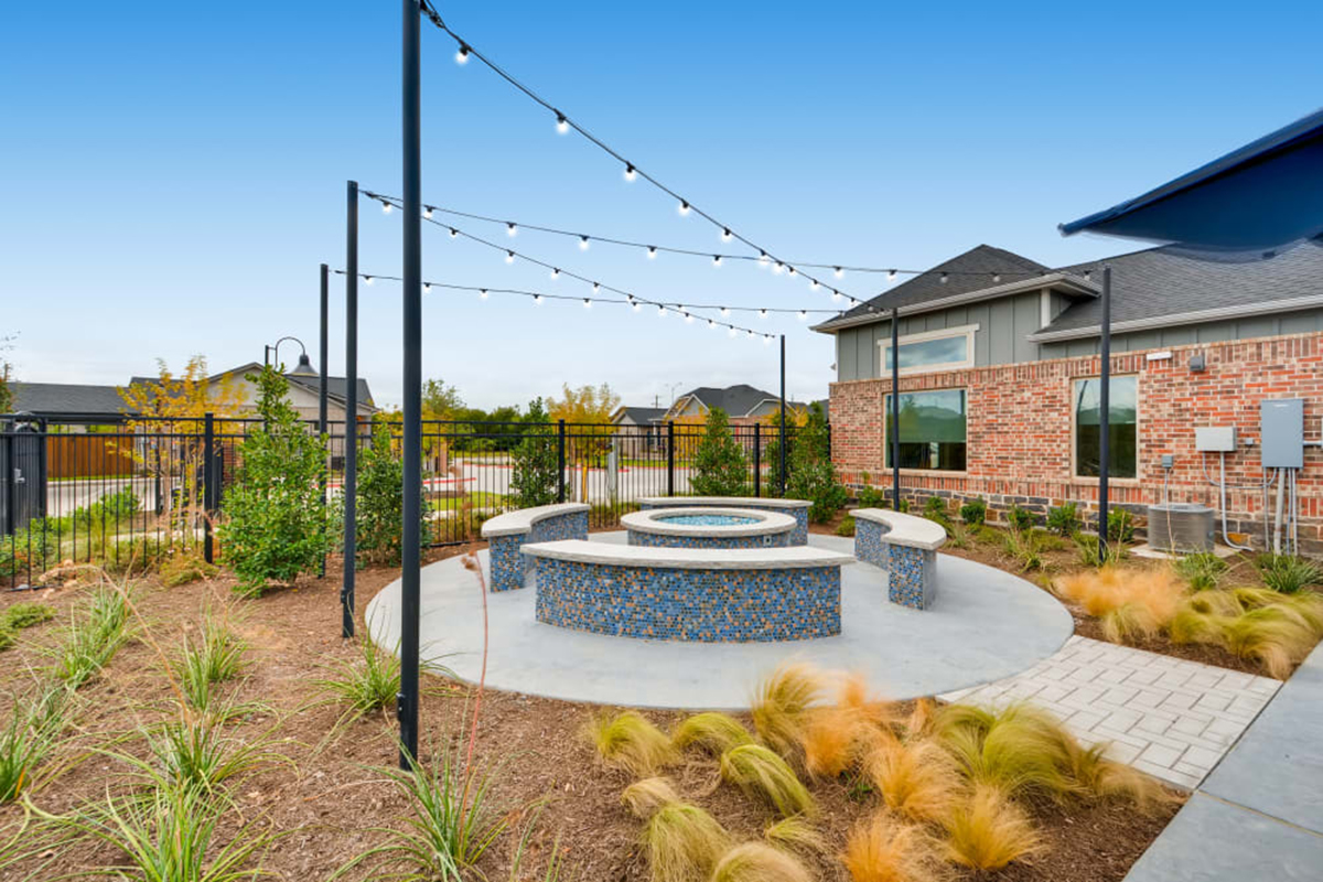 A circular outdoor fire pit with mosaic tile and curved benches, surrounded by plants, string lights above, next to a brick building on a clear day.