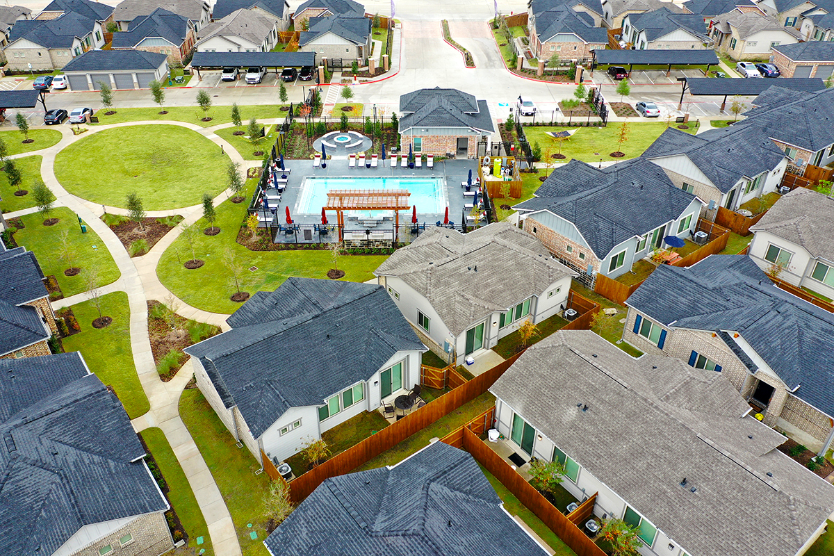 Aerial view of suburban neighborhood with houses, a circular green park area, paths, and a fenced community pool with seating and umbrellas at the center.