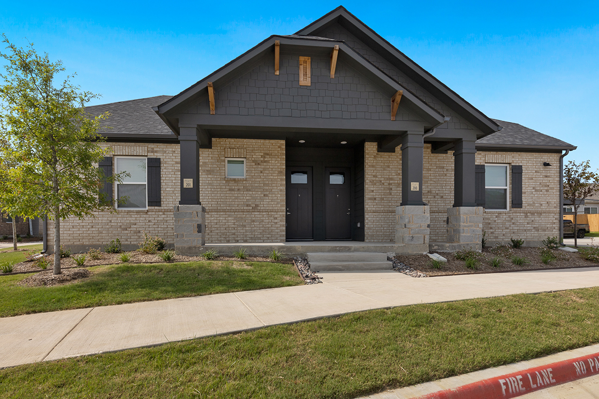 Single-story brick duplex with dark trim, two front doors, small covered porch, and minimal landscaping, set against a clear blue sky.