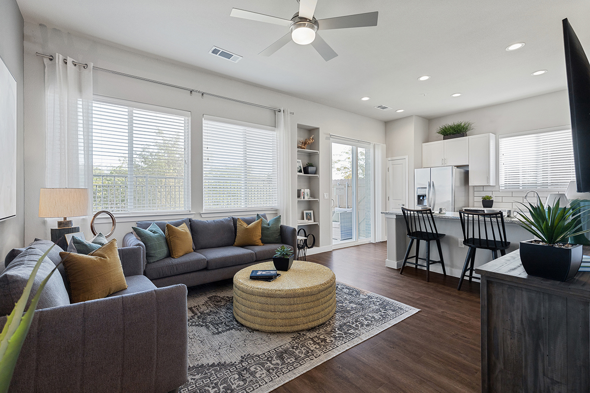 Modern living room with gray sofas, yellow pillows, round woven coffee table, rug, TV, and open kitchen with barstools. Large windows and glass doors provide natural light.