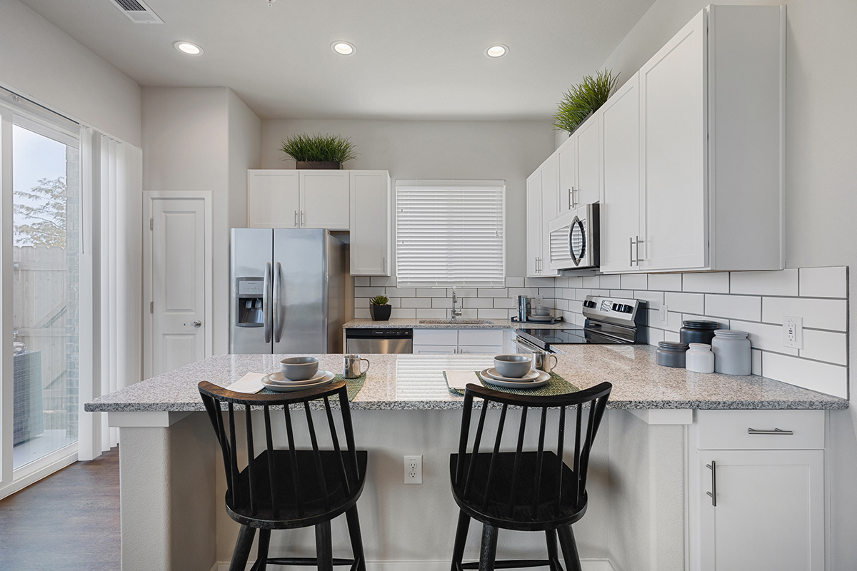 Modern kitchen with white cabinets, stainless steel appliances, subway tile backsplash, two black chairs at a gray countertop, and decorative plants on top of cabinets.